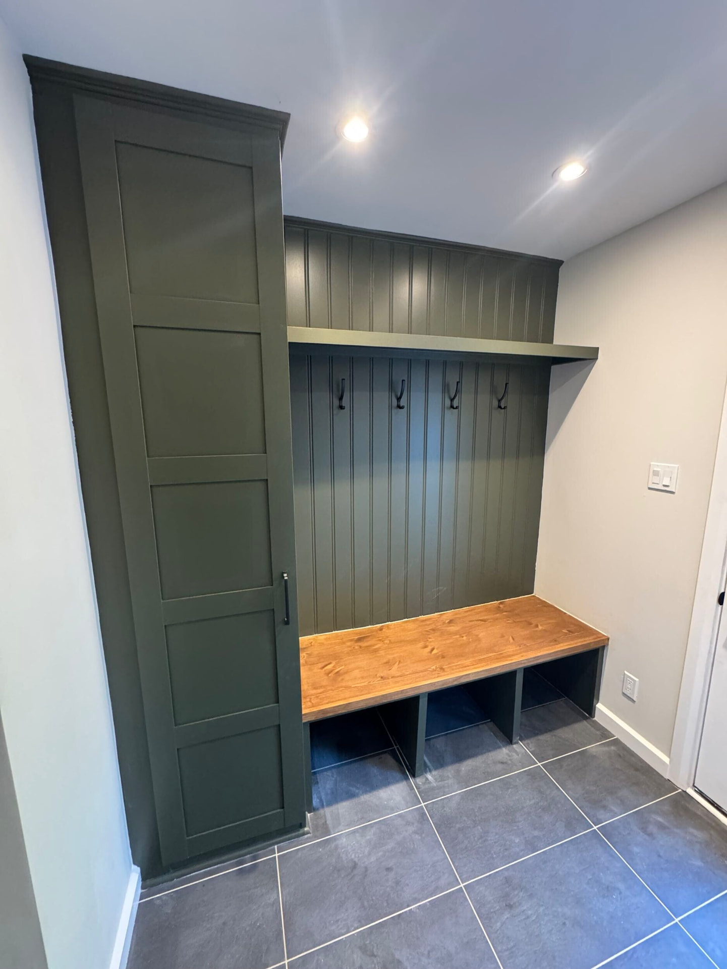 Custom olive green mudroom millwork with a pine bench, cubbies and black hooks over slate-look porcelain tile