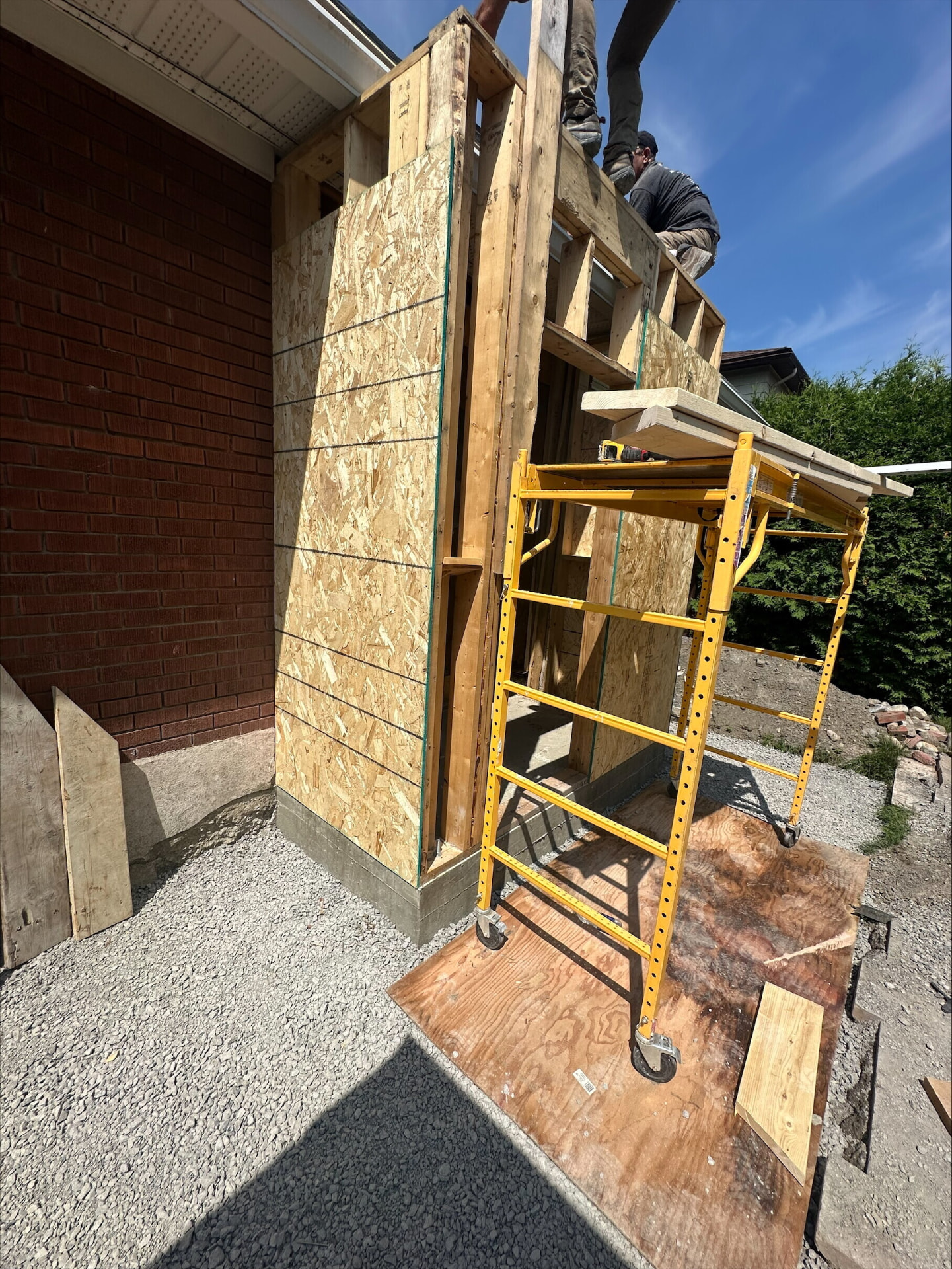 Framed and OSB-sheathed entrance addition with carpenters working from scaffolding beside a brick house