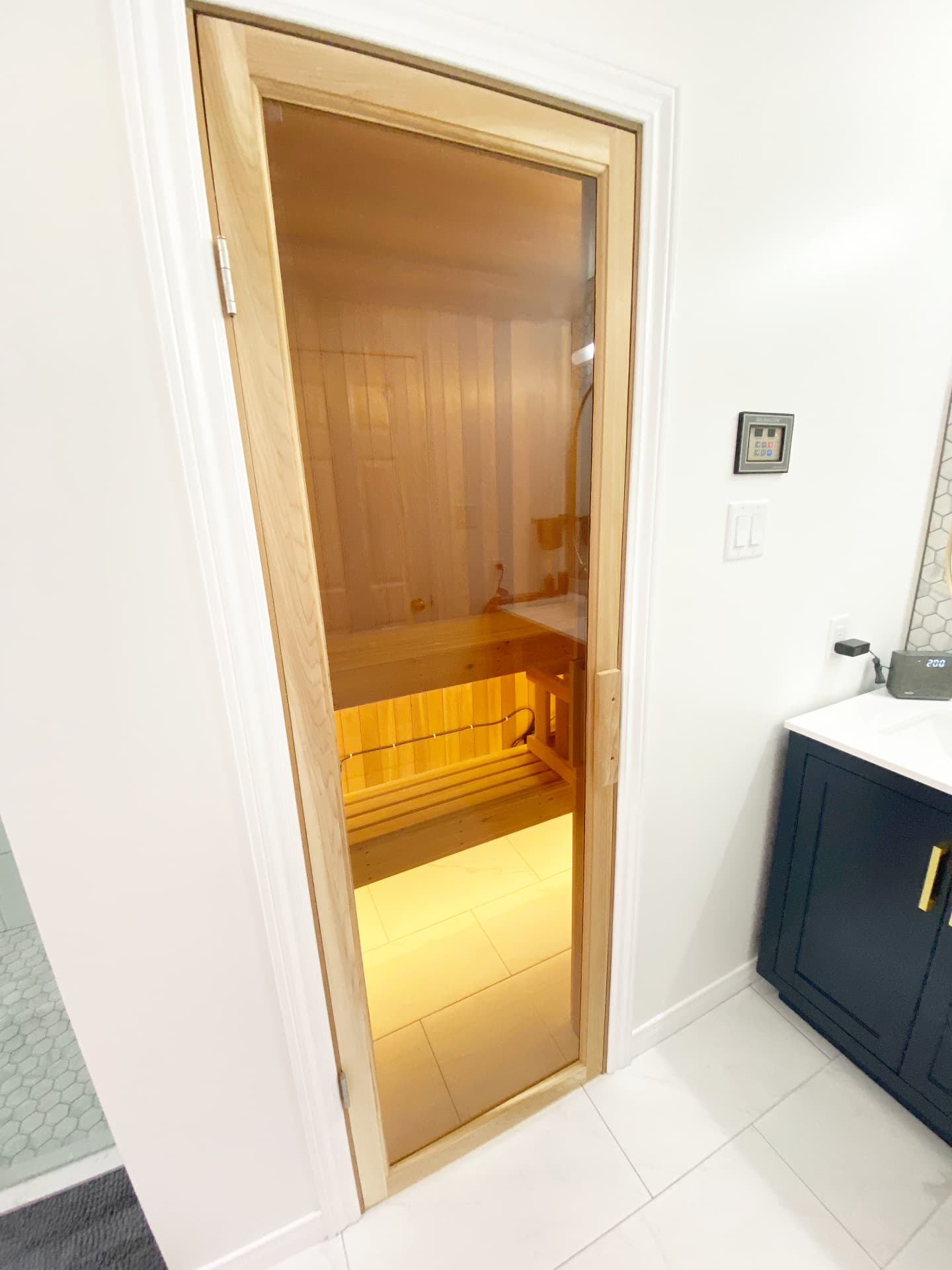 Cedar-framed bronze glass sauna door glowing beside a navy vanity in a white bathroom