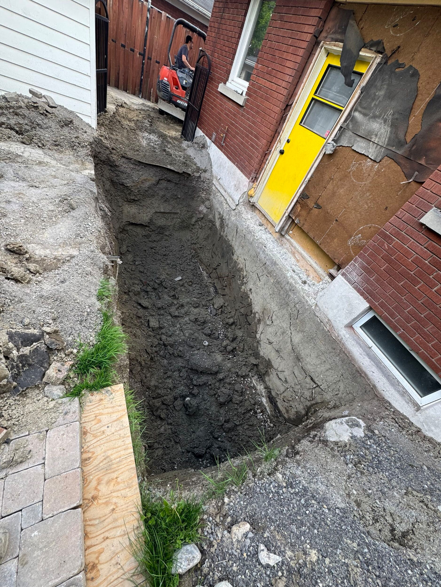 Excavated trench beside the foundation wall of a brick house with a mini excavator on the driveway