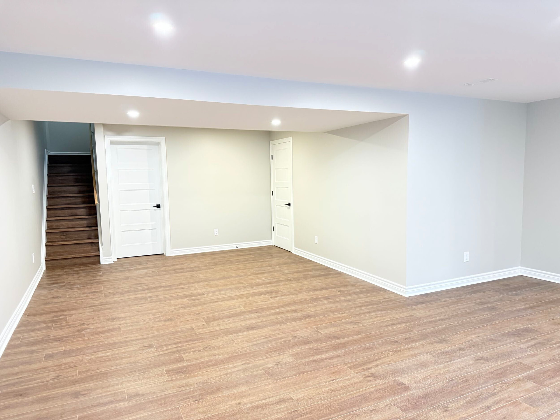 Finished basement room with wood-look plank flooring, white doors and the stair beyond
