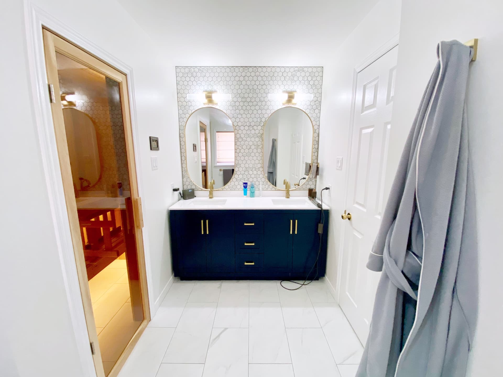 Wide view of the finished bathroom with marble floor tile, navy vanity and lit sauna doorway