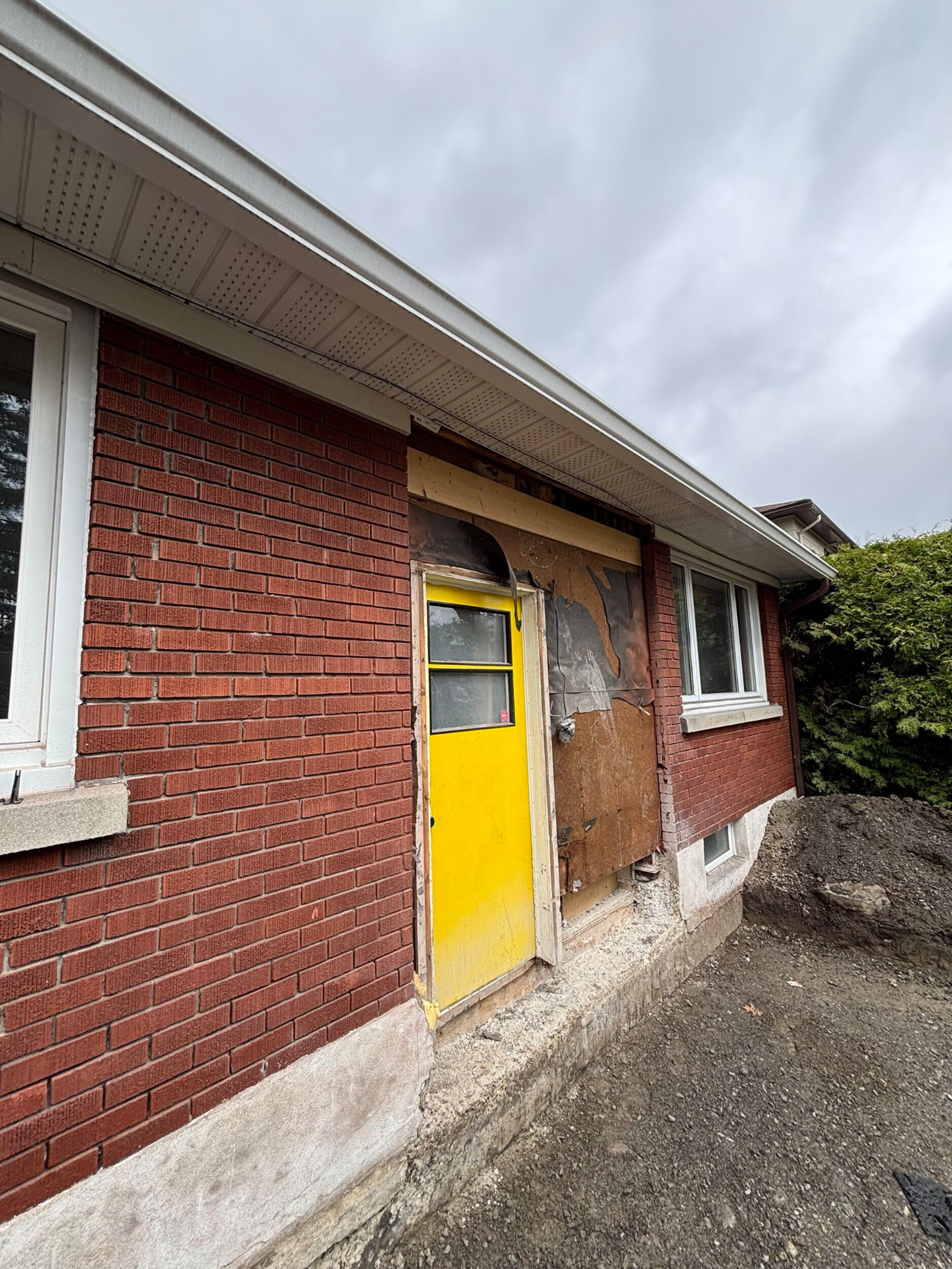 Original side entrance with a worn yellow steel door and exposed damaged sheathing on a brick bungalow