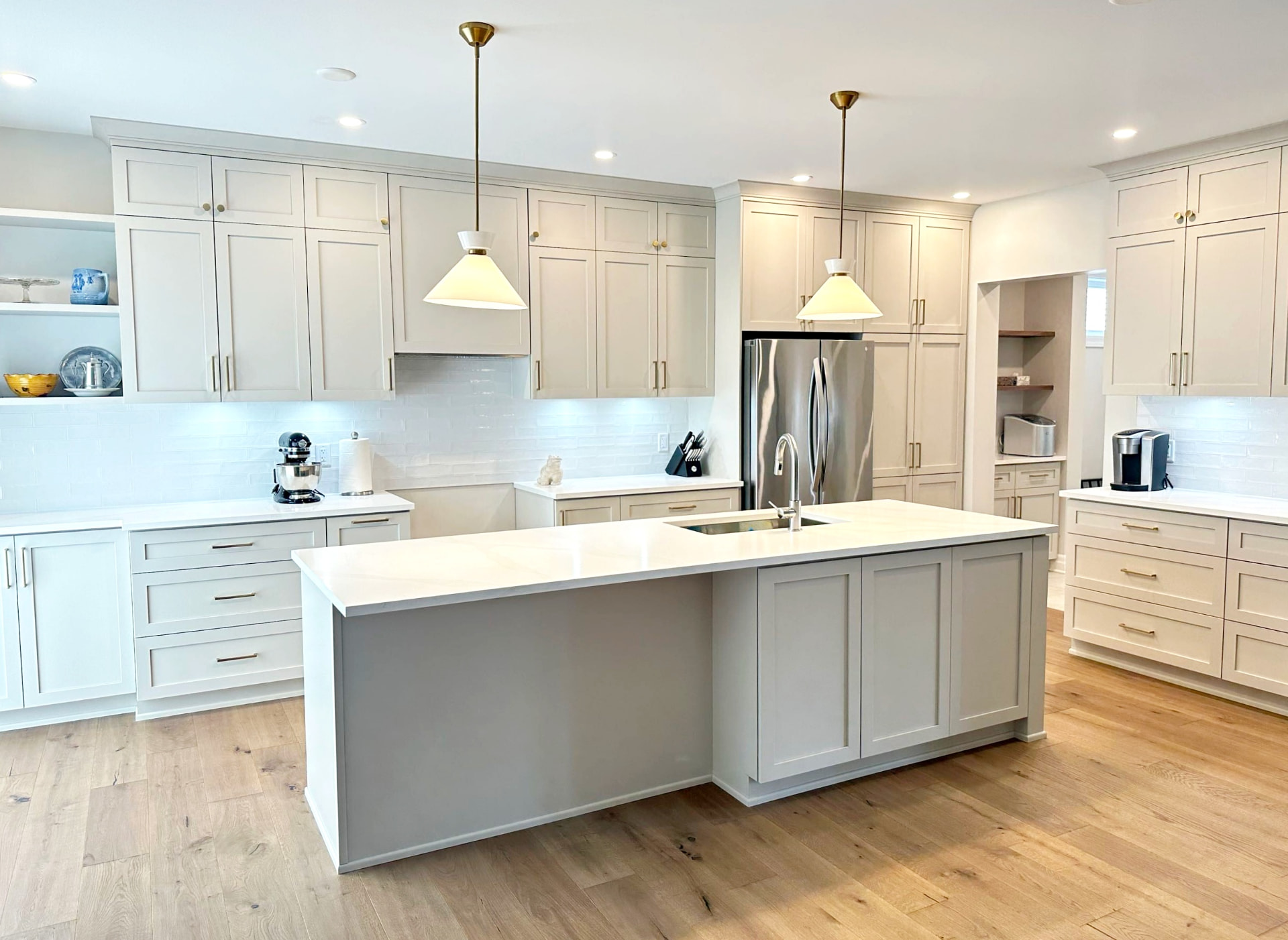 Wide view of the finished kitchen with pale shaker cabinetry, brass pendants and a quartz island